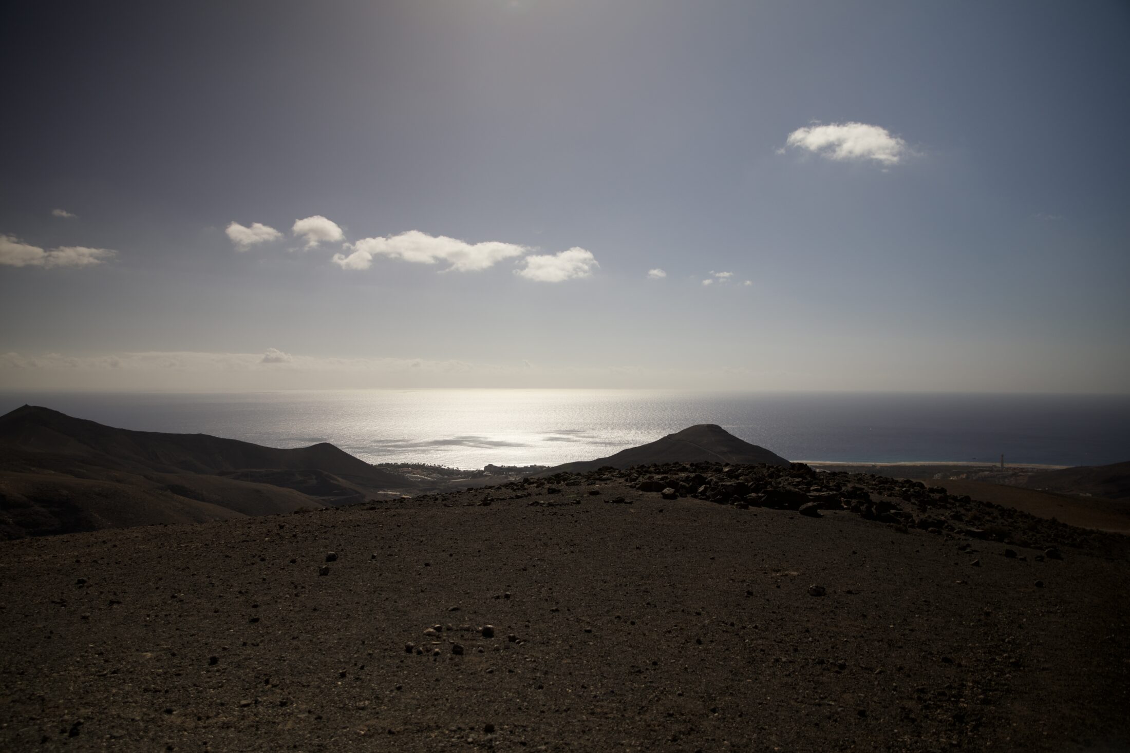 Die Sonne spiegelt sich im entfernten Meer, während ein paar Wolken am blauen Himmel zu sehen sind. Die Landmasse davor ist dunkel gegen das Licht.