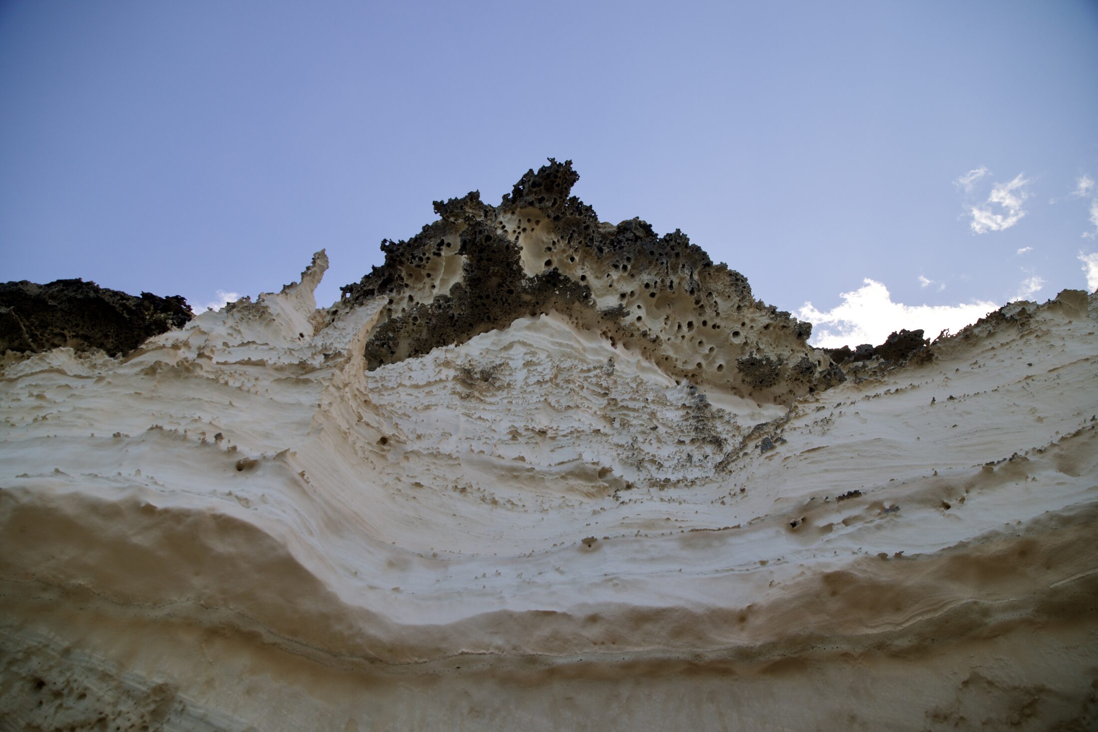 Blick einer steilen Klippe nach oben. Der Sandstein ist in Schichten, beige, hellbeige - fast weiß, und dunkelbraun. Bizarre Zacken sind im oberen dunkelbraunen Bereich zu sehen
