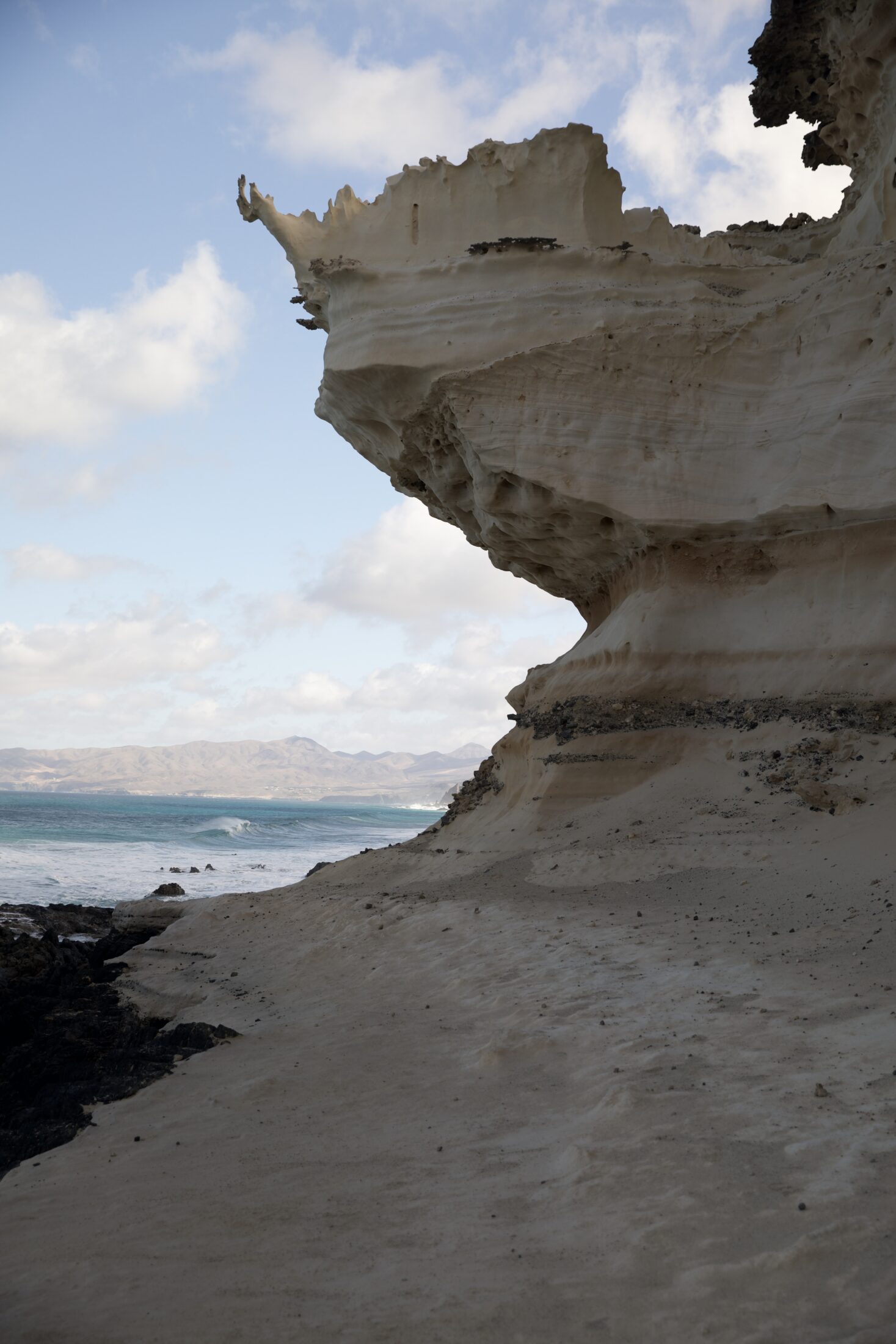 Der Sandstein ist in der Mitte so ausgewaschen, dass ein überhängender Teil an der Küste zu sehen ist. Im Hintergrund das Meer und ein weit entfernter Teil der Insel Fuertaventura