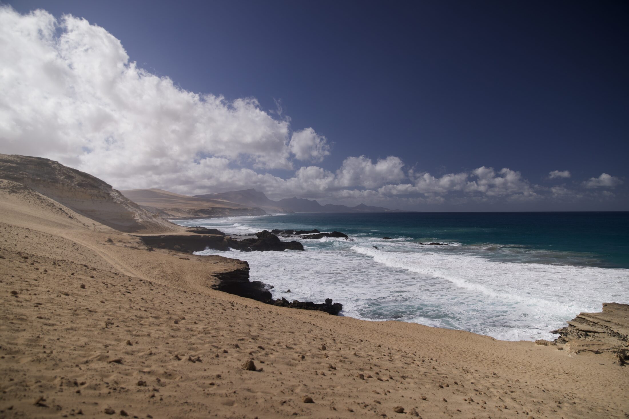 Blick entlang einer Küste. Das Meer ist weiß geschäumt in Küstennähe und tiefblau weiter weg. An der Küste ist kein Gewächs zu sehen.
