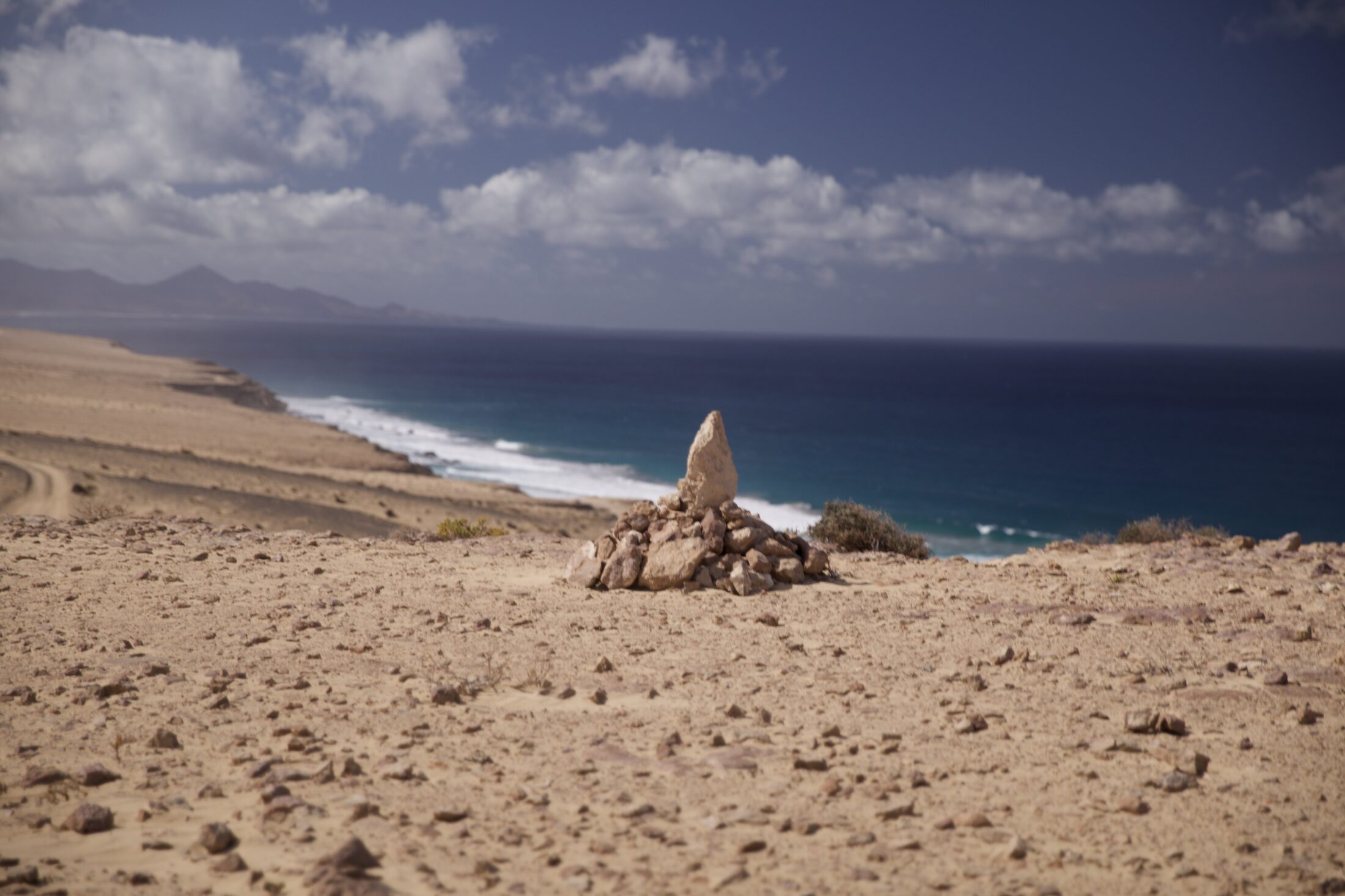 Ein großer oben spitzer Stein steht im Sand. Rundherum sind weitere Steine aufgeschichtet. Im Hintergrund sieht man Meer und Küstenlinie