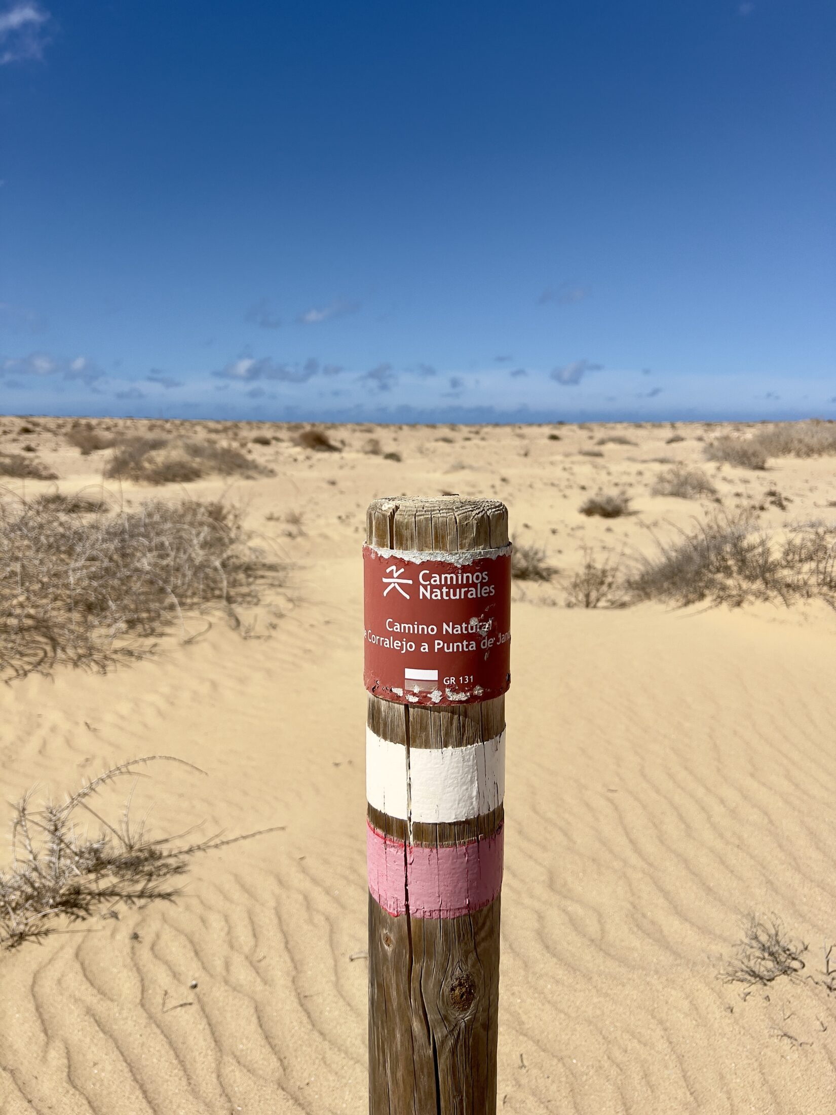 Ein Markierungspfahl für den Wanderweg GR 131 mitten im Sand. Ein rotes Schild darauf ist beschriftet mit "Camino Natural de Corralejo a Punta da Jandia" und "GR 131!. Darunter ist eine weiß-rote Markierung.