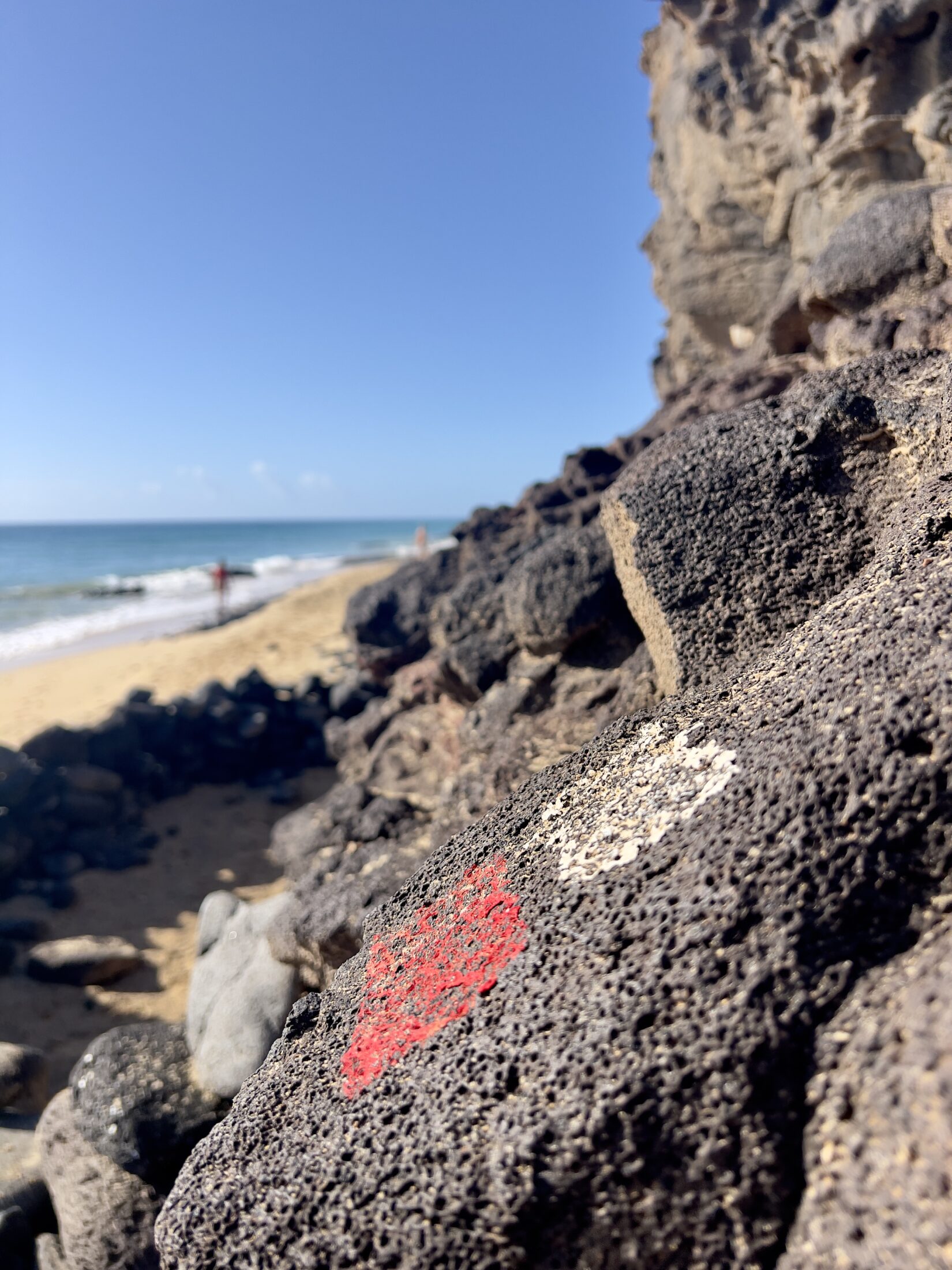 Eine weiß-rote Markierung auf einem Stein. Im Hintergrund ein Sandstrand in Unschärfe