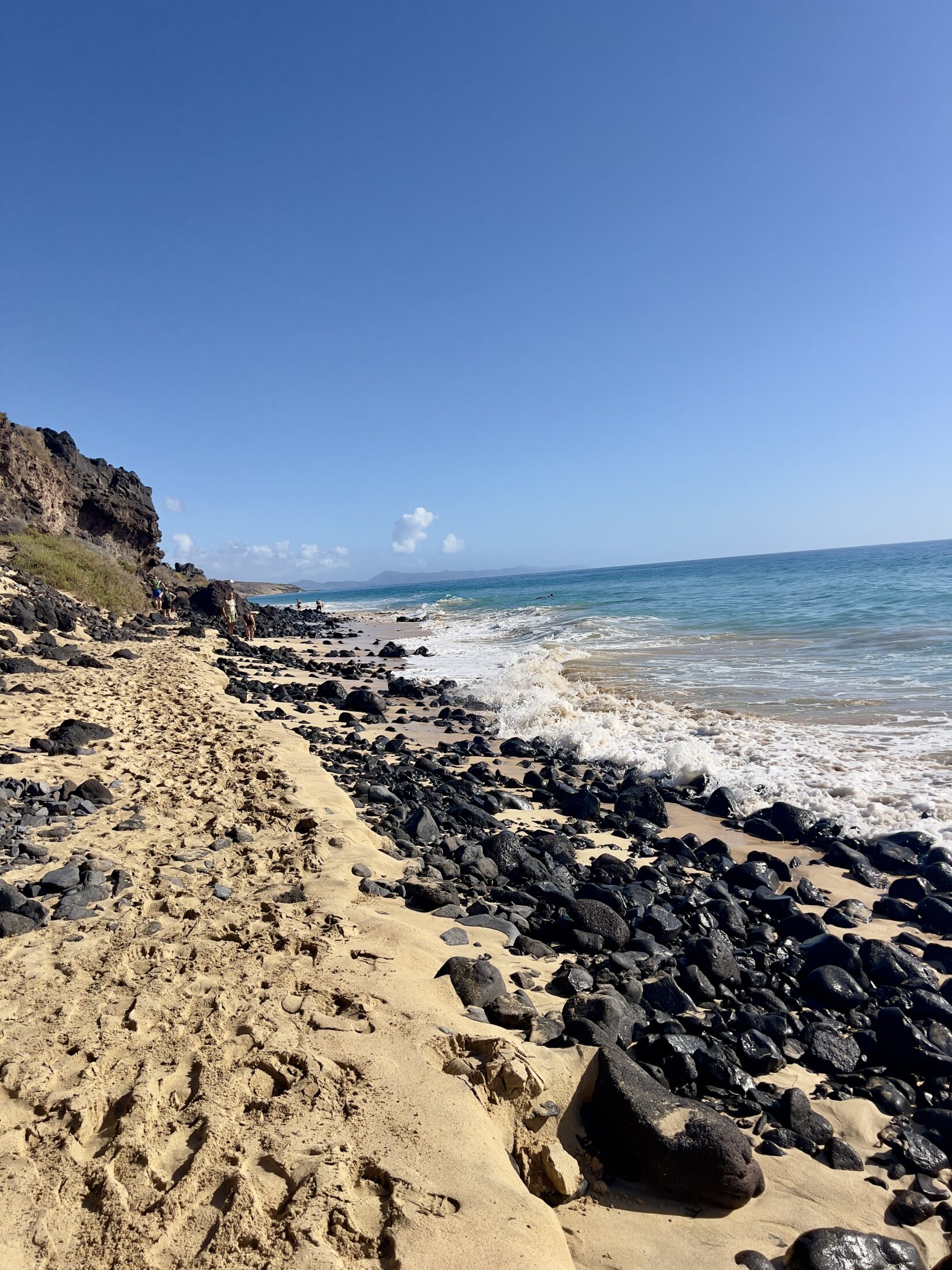 Vor den hohen Klippen ein Sandstrand und vielen schwarzen vulkanischen Gestein. Das Meer.