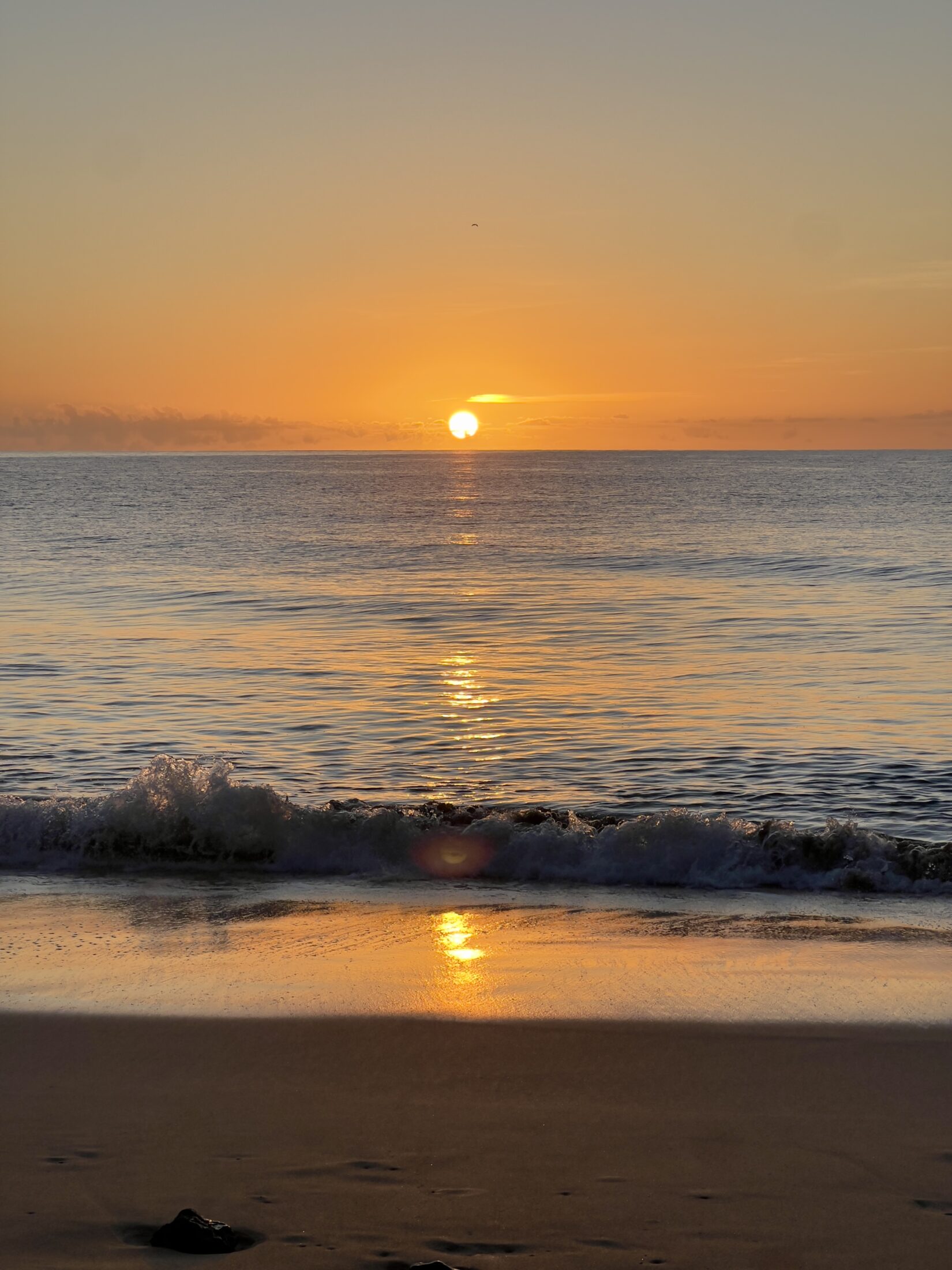 Die Sonne geht am Horizont über dem Meer auf. Eine Welle plätschert an den Sandstrand