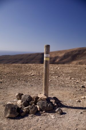 Eine hölzerne Wandermarkierung, oben weiß und gelb, steckt in wüstenartigen Boden. Rundherum sind Steine geschichtet.