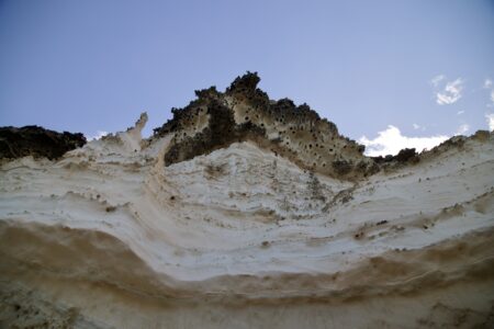 Blick einer steilen Klippe nach oben. Der Sandstein ist in Schichten, beige, hellbeige - fast weiß, und dunkelbraun. Bizarre Zacken sind im oberen dunkelbraunen Bereich zu sehen