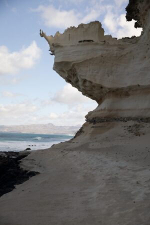 Der Sandstein ist in der Mitte so ausgewaschen, dass ein überhängender Teil an der Küste zu sehen ist. Im Hintergrund das Meer und ein weit entfernter Teil der Insel Fuertaventura
