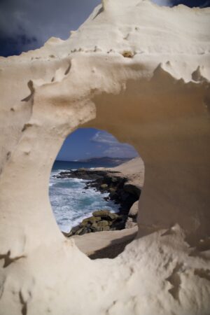 Durch ein Loch im Sandstein sieht man die dahinterliegende Küstenlinie.
