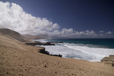 Blick entlang einer Küste. Das Meer ist weiß geschäumt in Küstennähe und tiefblau weiter weg. An der Küste ist kein Gewächs zu sehen.