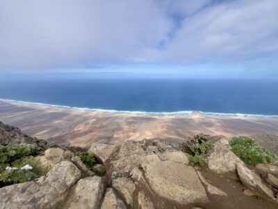 Blick von einem Berggipfel auf einen Sandstrand. Am Abgrund grünes niedriges Gebüsch und Steine