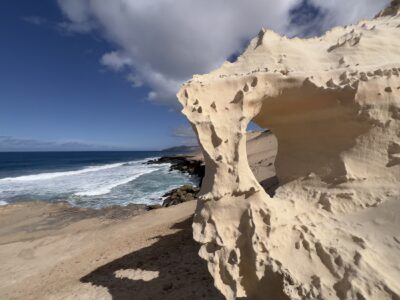 Meer und Küste im Hintergrund, im Vordergrund ein Sandstein mit einem Loch, durch das man sehen kann.