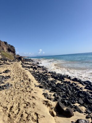 Vor den hohen Klippen ein Sandstrand und vielen schwarzen vulkanischen Gestein. Das Meer.