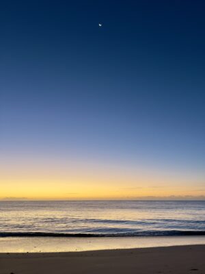 Meer mit Wellen und der Mond am Himmel. Der Horizont ist bereits orange kurz vor dem Sonnenaufgang